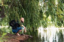 Side view of bald bearded male with backpack sitting on shore of lake under green tree on summer day — Stock Photo