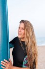 Vista laterale di surfista donna in piedi con bordo SUP blu sulla spiaggia sabbiosa in estate e guardando altrove — Foto stock