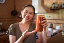 Весела етнічна домогосподарка показує скляні банки з домашнім томатним соусом маринари, сидячи за столом на кухні і дивлячись на камеру — стокове фото