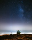 Далекий вид человека, стоящего на берегу моря возле спасательной башни под зрелищным ночным небом с светящимися звездами Млечного Пути — стоковое фото