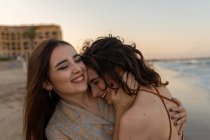 Jovens namoradas alegres abraçando uns aos outros enquanto estão de pé na praia de areia perto do mar acenando ao pôr do sol — Fotografia de Stock