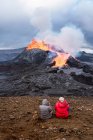 Vista posterior de viajeros irreconocibles admirando Fagradalsfjall con fuego y lava mientras toma fotos y se sienta en el monte en Islandia - foto de stock