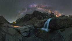 Анонимный путешественник с факелом, созерцающий водопад, струящийся среди неровной скалистой местности под ночным звездным небом с ярким светящимся Млечным Путем — стоковое фото