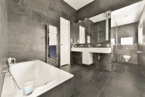 Інтер'єр просторої ванної кімнати з сірими плитковими стінами і підлогою, білою ванною і раковинами — стокове фото