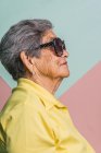 Vista laterale di felice donna moderna invecchiata con i capelli grigi e in occhiali da sole alla moda su sfondo rosa in studio e guardando altrove — Foto stock