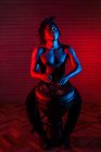 Мечтательный черный музыкант с обнаженным туловищем, играющий на африканском барабане в студии с красным и синим неоновым светом — стоковое фото