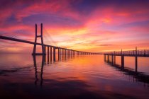 Силуэт моста Васко да Гама и длинная набережная, расположенная на спокойной реке Лус на фоне облачного закатного неба вечером в Лисбоне, Португалия — стоковое фото