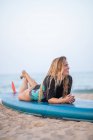 Вид сбоку на позитивную женщину-серфингистку в купальниках, лежащую на доске на песчаном берегу на фоне моря и отводящую взгляд — стоковое фото
