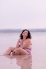 Délicieuse femelle courbée en bikini assise dans l'eau de l'étang rose en été et regardant la caméra — Photo de stock