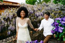 Contenu couple multiracial tenant la main tout en marchant dans le jardin avec des fleurs de glycine pourpre en fleurs et profiter week-end ensemble — Photo de stock