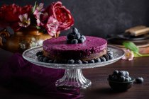 Délicieux gâteau mousse myrtille avec crème pourpre décorée de baies fraîches servi sur support en verre sur table sombre avec des fleurs — Photo de stock