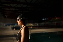 Молода красива жінка, що сидить на краю критого басейну з чорним купальником, дивиться на камеру — стокове фото