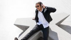 Высокий угол молодого этнического мужчины с плетеными волосами, одетого в стильный костюм и солнцезащитные очки, наслаждающегося музыкой через беспроводные наушники во время отдыха на солнечной городской площади — стоковое фото