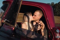 Jeune couple amoureux assis dans un pick-up vintage rouge et embrasser au coucher du soleil en été — Photo de stock