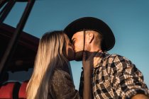 De baixo vista lateral da jovem mulher amorosa beijando o homem em chapéu de cowboy ternamente perto do carro no fundo do céu azul à noite — Fotografia de Stock