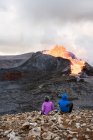 Обратный вид на неузнаваемых путешественников, любующихся огнем и лавой на Фаградальсфьоле, сидящих на горе в Исландии — стоковое фото