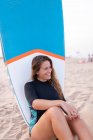 Cheerful female surfer sitting with blue SUP board on sandy seashore in summer and looking away — Stock Photo