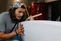 Konzentrierter männlicher Hipster mit Schreibwerkzeug und Bleistift beim Markieren des Surfbretts vor dem Formen in der Werkstatt — Stockfoto