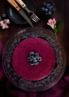 Смачний чорничний мус з фіолетовим кремом, прикрашений свіжими ягодами, які подаються на скляній підставці на темному столі з квітами — стокове фото