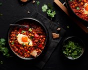Top view of chakchouka with sunny side up egg in delicious tomato sauce with rye bread piece in bowl between fresh cilantro on dark background — Stock Photo