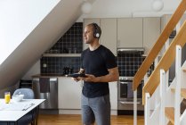 Zufriedener Mann mit Kopfhörer, der Lieder hört und Tablet benutzt, während er zu Hause beim Frühstück in der Küche steht und wegschaut — Stockfoto