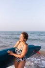 Mujer en traje de baño de pie con tabla SUP en agua de mar en verano y mirando hacia otro lado - foto de stock