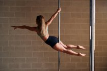 Side view of graceful female dancer hanging with outstretched arm on pole while rehearsing in modern studio — Stock Photo