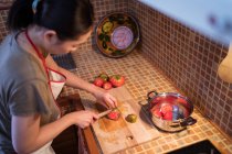 Вид зверху на етнічну жінку в фартусі, що розрізає стиглі помідори на дошці під час приготування обіду на кухні вдома — стокове фото