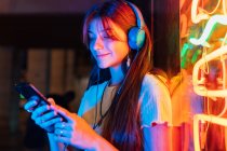 Cheerful young female with cellphone listening to song from headphones against colorful neon lights in evening town — Stock Photo