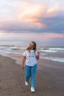 Positive young female in trendy sunglasses and stylish outfit walking at seaside against sea in summer evening — Stock Photo
