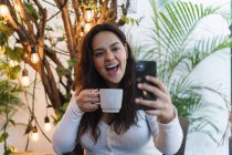 Alegre joven latinoamericana hembra tomando selfie en el teléfono móvil mientras bebe café en la cafetería con plantas verdes en el fondo - foto de stock