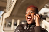 Вид збоку красива чорна афро-жінка розмовляє зі своїм смартфоном, посміхаючись з розмитим фоном на сонячний день — стокове фото