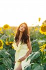 Gracieuse jeune femme hispanique en robe jaune élégante debout au milieu de tournesols en fleurs dans le champ de campagne dans la journée ensoleillée d'été en regardant la caméra — Photo de stock