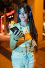 Interested female with beverage chatting on cellphone while listening to music from headset in night city — Stock Photo