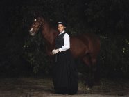 Senhora adulta afro-americana confiante em roupas elegantes e chapéu de pé com cavalo marrom enquanto olha para a câmera perto de árvores durante o dia — Fotografia de Stock