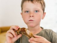 Criança adorável comendo costelas de porco apetitosas com boca cheia durante o almoço em casa e olhando para longe — Fotografia de Stock