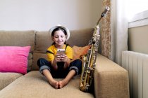 Criança em chapéu mensagens de texto no celular enquanto sentado no sofá com saxofone na sala de estar — Fotografia de Stock
