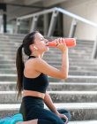 Young sportswoman in black activewear listening to music and sipping fresh water with closed eyes while sitting on ground during break in fitness training — Stock Photo