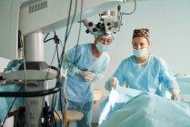 Médica atenta em uniforme estéril contra colega de trabalho desviando o olhar enquanto se prepara para cirurgia no hospital com microscópio — Fotografia de Stock
