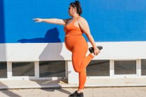 Vista lateral de mujer atleta regordeta en ropa deportiva haciendo ejercicio en la pasarela de baldosas en la ciudad soleada - foto de stock