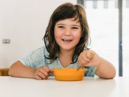 Entzückendes Kind isst leckere Sahnesuppe aus Plastikschüssel, während es zu Hause zu Mittag am Tisch sitzt — Stockfoto