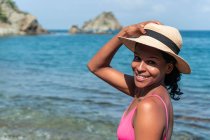 Vista lateral del alegre turista étnico femenino en traje de baño y sombrero mirando a la cámara en la costa del océano - foto de stock