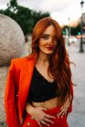Charming female with long red hair and in trendy orange suit standing in street in evening and looking at camera — Stock Photo