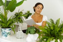 Pensive ethnic female with pen and agenda between green plants in pots in house garden — Stock Photo