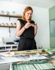 Веселая женщина-дизайнер фотографирует картины на смартфоне, стоя рядом со столом и работая в творческом рабочем пространстве — стоковое фото