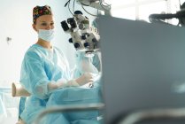 Médecin adulte attentif féminin en masque jetable et casquette ornementale travaillant en clinique avec microscope chirurgical — Photo de stock