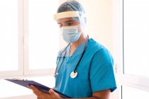 Médecin masculin concentré avec stéthoscope et masque lisant le rapport médical sur presse-papiers pendant qu'il se tenait debout à la clinique — Photo de stock