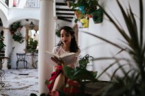 Contenido étnico femenino en traje elegante sentado en el taburete en el patio y la lectura de la novela en el libro mientras disfruta del fin de semana de verano - foto de stock
