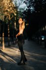 Seitenansicht einer glücklichen Frau in trendiger Kleidung, die an sonnigen Tagen in der Stadt steht und in die Kamera blickt — Stockfoto