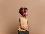 Satisfied preteen boy in red headphones listening to music and signing song on brown background in studio — Stock Photo